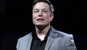 Elon Musk pe scenă, îmbrăcat într-un costum gri, cu o cămașă neagră, pe fundal negru. Elon Musk vrea să se implice într-o nouă industrie