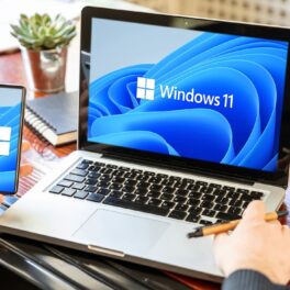 Utilizator care se află în fața unui laptop și cu un telefon în mână, ambele cu Windows 11 pe ecran. Microsoft a confirmat versiunea 22H2 a Windows 11