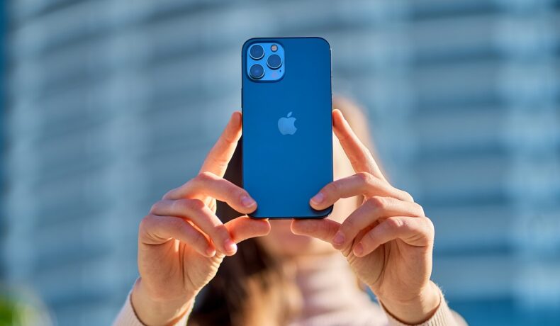 Utilizatoare care ține în mână un telefon iPhone 12, de culoare albastră, cu o clădire pe fundal. iPhone 14 Pro ar putea avea o cameră de 48 MP