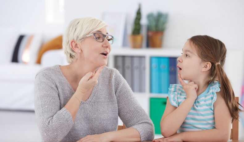 Femei în vârstă care învață un copil să vorbească, ambii stau la o masă și își ating gâtul. Mulți oameni se întreabă ce e afazia: cauze și simptome