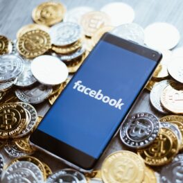 Telefon negru, cu Facebook pe ecran, care stă pe un maldăr de monede, fundal gri. Meta ar crea „Zuck Bucks”, potrivit unui raport