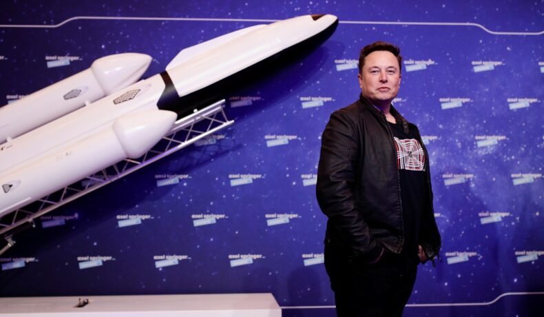 Elon Musk, pe covorul roșu la Axel Springer Award, 2020. Îmbrăcat în negru, pe fundal albastru, cu o rachetă. Recent, Starlink a blocat un atac electromagnetic în Ucraina
