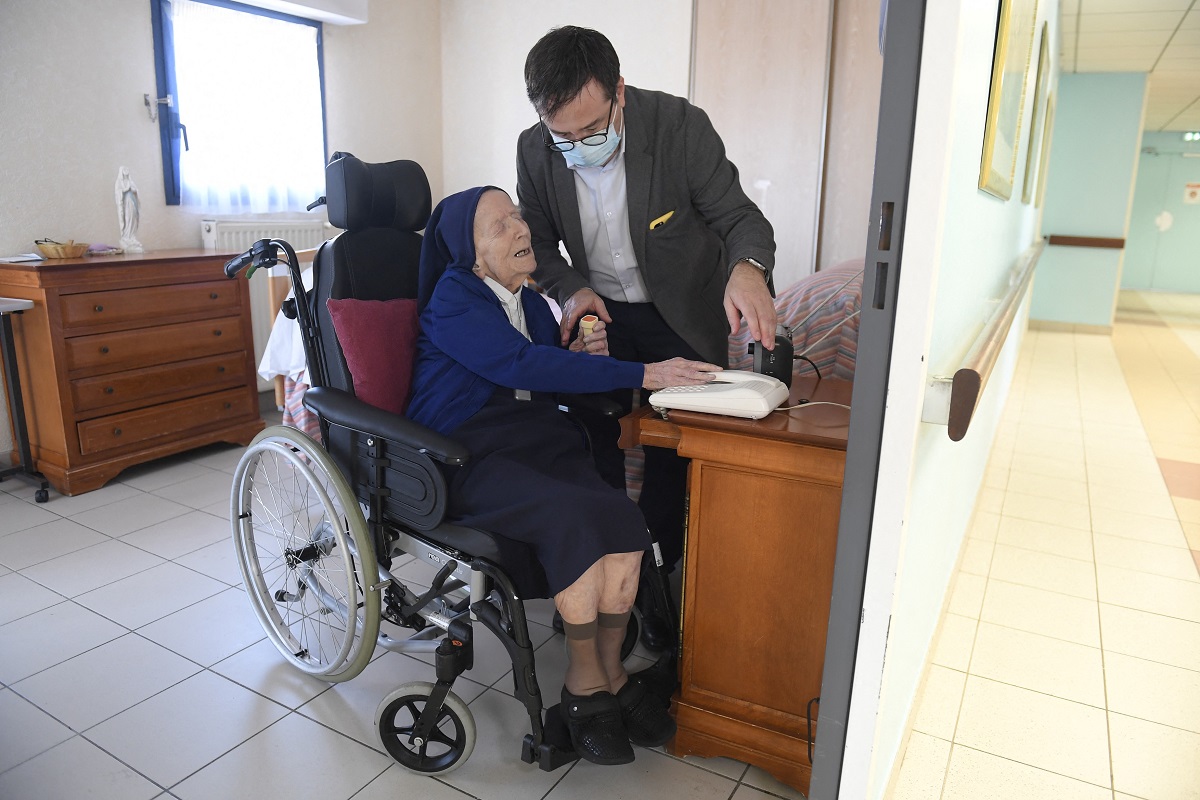 Sora André, Lucile Randon, în scaunul cu rotile, într-o clădire, ajutată de un bărbat