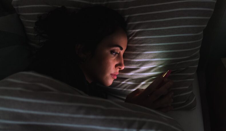 Femeie care stă în pat și se uită în telefon. Experții au dezvăluit recent de ce nu trebuie să folosești telefonul în pat