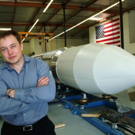 Elon Musk lângă o rachetă albă și steagul american pe fundal. Recent, Elon Musk ar fi fost ridiculizat într-o reclamă Ford