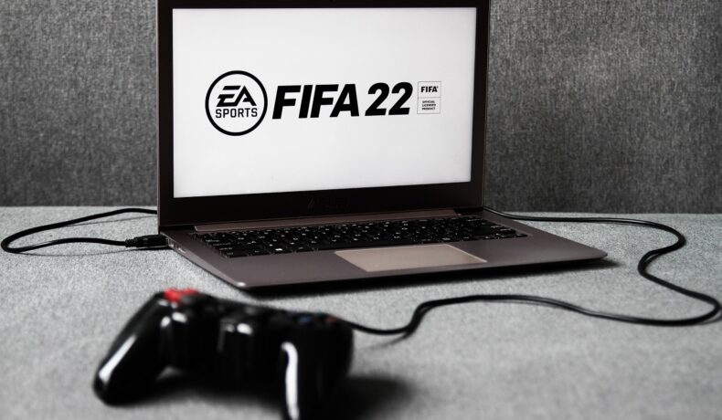 Laptop negru cu FIFA 22 pe ecranul alb, cu fundal gri. FIFA 22 include un echipament cu motive românești