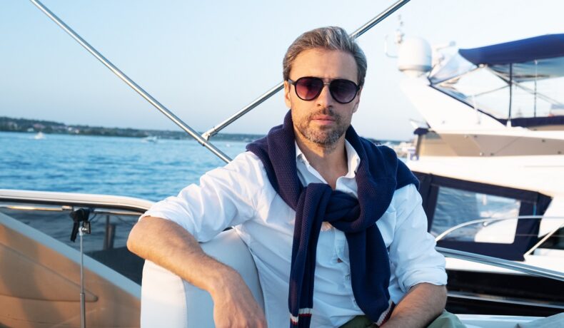 Miliardar care stă pe un iaht, cu marea pe fundal și o bluză albă. SUA e țara cu cei mai mulți miliardari