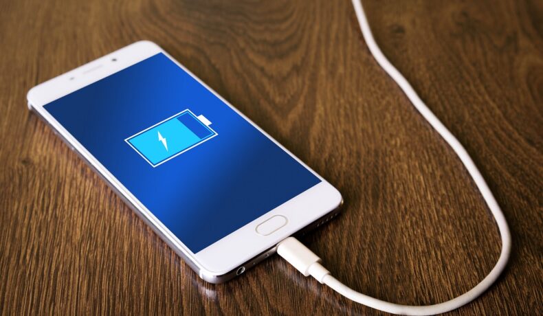 Telefon alb, cu ecran albastru, care e pe lemn închis la culoare, la încărcat. Mulți s-au întrebat de ce nu au telefoanele baterii mai mari