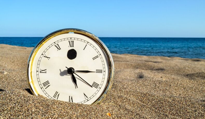 Ceas de metal în nisip, pe o plajă, cu cerul albastru și marea pe fundal. Experții au dezvăluit de ce oscilează durata unei zile la fiecare 6 ani