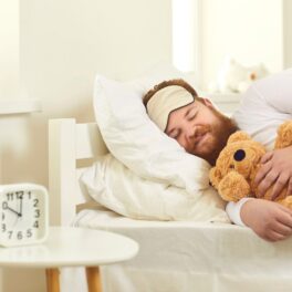 Bărbat cu barbă roșcată care doarme cu un ursuleț în brațe. Experții cred că o nouă tehnologie poate să ajute oamenii să slăbească în timp ce dorm
