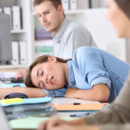 Femeie care a adormit la birou, la care se uită îngrijorați colegii. Cauza narcolepsiei a fost descoperită recent