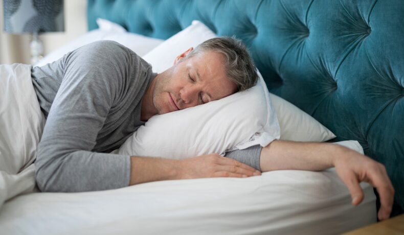 Bărbat care doarme în pat, cearșafuri albe, tăblie albastră. Experții au dezvăluit ce ți se poate întâmpla dacă dormi mai puțin de 5 ore pe noapte