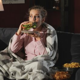 Femeie îmbrăcată în pijama care stă pe canapea și mănâncă un sandwich. Experții au dezvăluit recent de ce nu trebuie să mănânci seara târziu