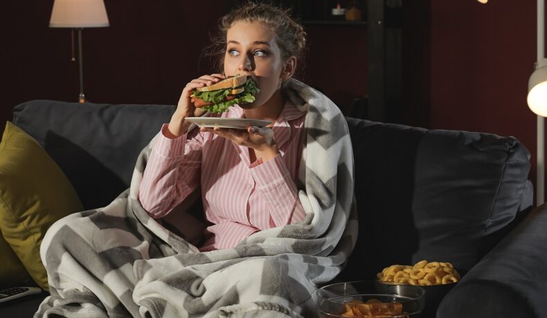 Femeie îmbrăcată în pijama care stă pe canapea și mănâncă un sandwich. Experții au dezvăluit recent de ce nu trebuie să mănânci seara târziu