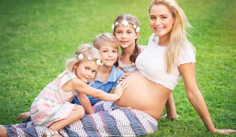 Femeie însărcinată care stă pe iarbă, cu 3 copii care o ating pe burtică. Experții au dezvăluit câți copii poți avea