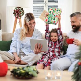 Familie care stă pe canapea și deschide cadouri, inclusiv o tabletă albă, care nu se numără printre acele gadget-uri smart pentru casă