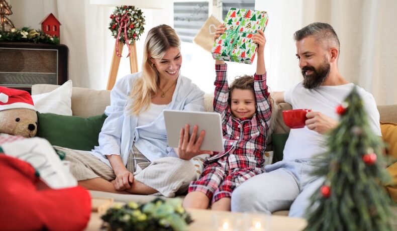 Familie care stă pe canapea și deschide cadouri, inclusiv o tabletă albă, care nu se numără printre acele gadget-uri smart pentru casă