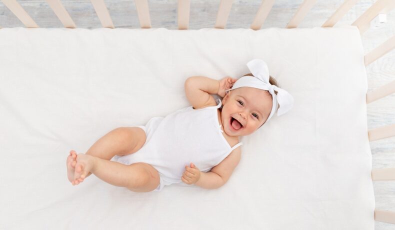 Bebeluș îmbrăcat în alb, care stă într-un pat alb și care are și o fundă albă pe cap. Într-un studiu recent, 10.000 de bebelusi vor avea genomurile secvențiate