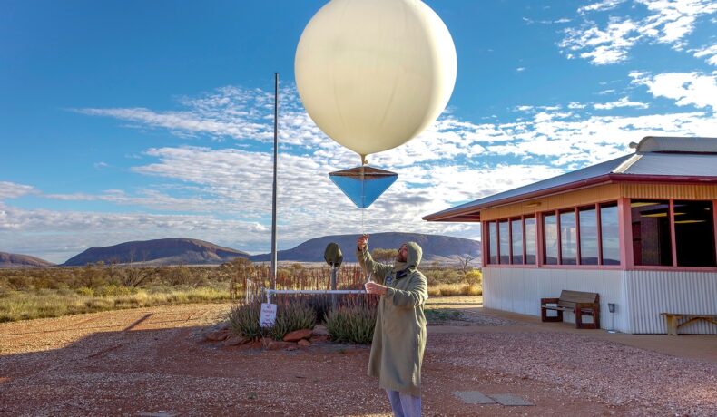 Bărbat care lansează un balon meteo alb în aer, lângă o stație meteo. O companie a început să lanseze particule în aer