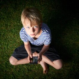 Băiat blond, care stă în iarbă, cu un telefon în mână. Experții au dezvăluit ce efect poate avea comunicarea prin intermediul tehnologiei asupra copiilor