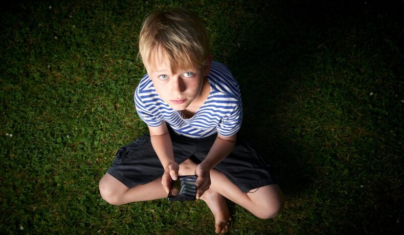 Băiat blond, care stă în iarbă, cu un telefon în mână. Experții au dezvăluit ce efect poate avea comunicarea prin intermediul tehnologiei asupra copiilor