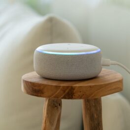 Boză Amazon Echo albă, pe o masă de lemn, lângă o canapea albă. Experții recomandă să nu ții boxa Alexa în dormitor