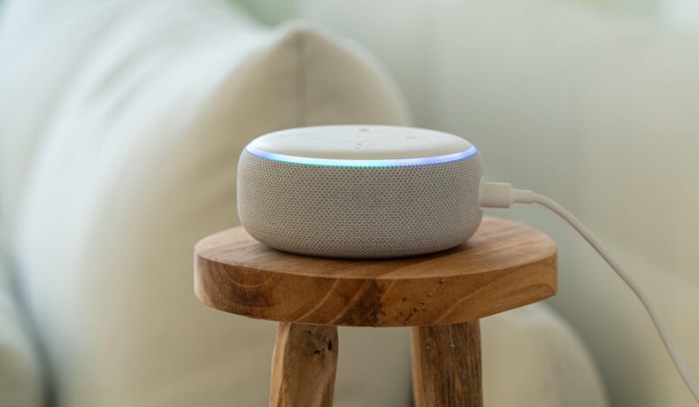 Boză Amazon Echo albă, pe o masă de lemn, lângă o canapea albă. Experții recomandă să nu ții boxa Alexa în dormitor
