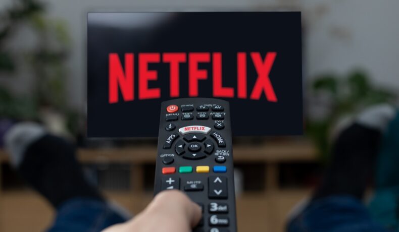 Persoană care are o telecomandă în mână și se uită la un televizor cu Netflix pe el. Netflix introduce o schimbare importantă în 2023
