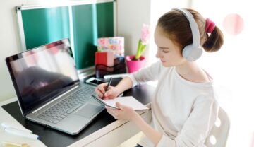 Elevă care învață la birou, în fața unui laptop, cu căști pe ureche. ChatGPT e programul care poate face temele elevilor