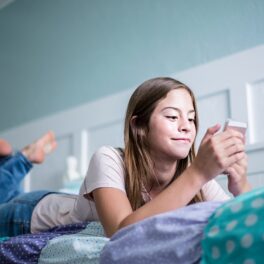 Fată adolescentă care stă în pat în cameră albastră și care se uită la un telefon mobil. Adolescenții din SUA refuză să folosească telefoane Android