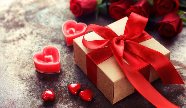 Cutie de cadou cu o fundă rpșie, pe fundal gri, înconjurată de lumănări în formă de inimă. Cadouri pentru Valentine's Day pot fi și tech