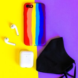 Telefon mobil în culori de curcubeu, pe fundal cu mov și galben, cu căști wireless albe și o mască neagră lângă. O nouă tehnologie explică cum poți schimba culoarea telefonului