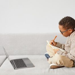 Băiat care stă pe canapea, scrie într-un carnet, și are un laptop în față. Similar cu David Balogun, unul dintre cei mai tineri absolvenți de liceu din lume