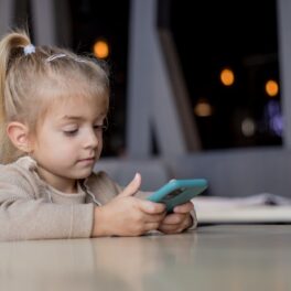 Fetiță blondă cu părul prins, care are un telefon albastru în mână și stă la o masă bej. Experții au dezvăluit până la ce vârstă nu ar trebui copiii să folosească rețelele de socializare