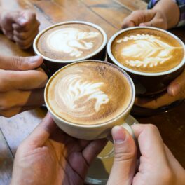 Oameni care țin 3 cești de cafea în mâini, deasupra unei mese din lemn, pentru a ilustra ce efecte poate avea cofeina în corp