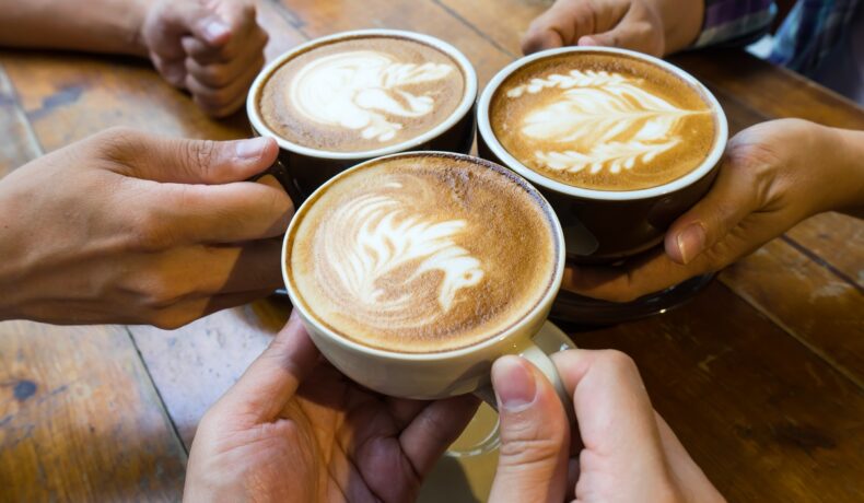 Oameni care țin 3 cești de cafea în mâini, deasupra unei mese din lemn, pentru a ilustra ce efecte poate avea cofeina în corp