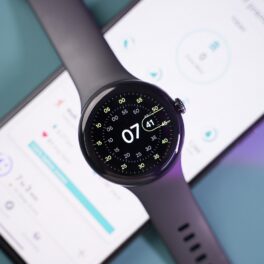 Pixel Watch negru, deasupra unui telefon aprins, pe fundal gri cu mov. Recent, a fost dezvăluit costul de producție pentru Pixel Watch