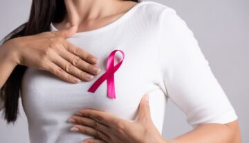 Femeie îmbrăcată în alb, care își palpează sânul, cu fundiță roz, pentru a ilustra cum experții au putut detecta un caz de cancer mamar