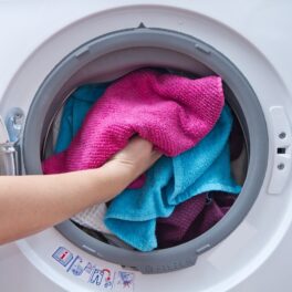 Mână care intră într-o mașinnă de spălat plină de prosoape colorate, roz și albastru. O substanță folosită pentru a curăța hainele poate fi cauză principală pentru Parkinson