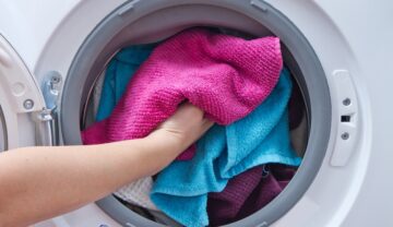 Mână care intră într-o mașinnă de spălat plină de prosoape colorate, roz și albastru. O substanță folosită pentru a curăța hainele poate fi cauză principală pentru Parkinson