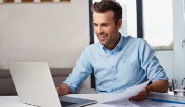 Bărbat îmbrăcat în cămașă albastră, la birou, în fața unui laptop, care zâmbește. Experții au dezvăluit țările cu cel mai ieftin Internet din lume