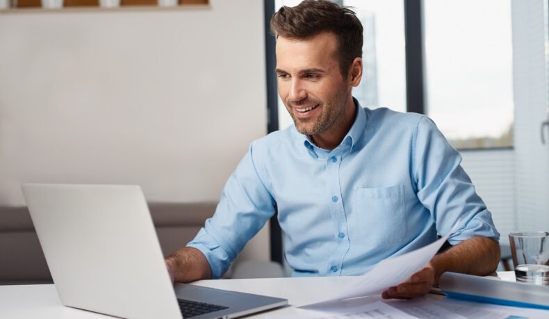Bărbat îmbrăcat în cămașă albastră, la birou, în fața unui laptop, care zâmbește. Experții au dezvăluit țările cu cel mai ieftin Internet din lume