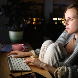 Femeie blondă care stă în fața unui calculator noaptea, cu genunchii la piept, pentru a ilustra care e diferența dintre persoanele matinale și cele nocturne