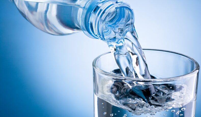 Sticlă de apă care e scursă într-un pahar transparent, pe fundal albastru, pentru a ilustra cel mai rapid mod de a goli o sticlă cu apă
