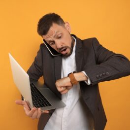 Bărbat îmbrăcat în costum care ține un laptop în mână și se uită la ceas, pe fundal portocaliu, pentru a ilustra de ce întârzie unii oameni mereu