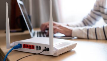 Router alb, care stă pe birou, lână un utilizator care scrie la laptop, pentru a ilustra secretele care se pot ascunde într-un router
