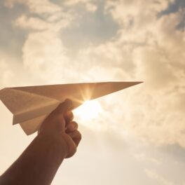 Persoană care ține un avion de hârtie în mână, cu cerul pe fundal