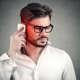 Bărbat cu ochelari, în cămașă albă, pe frundal gri, care vorbește la telefon, pentru a ilustra ce efect au telefoanele asupra creierului