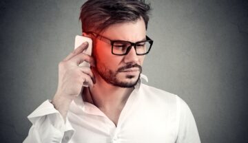 Bărbat cu ochelari, în cămașă albă, pe frundal gri, care vorbește la telefon, pentru a ilustra ce efect au telefoanele asupra creierului