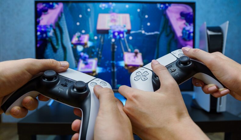 2 utilizatori cu controllere în mână, care joacă PlayStation, în fața unui monitor, pentru a ilustra cele mai bune jocuri PlayStation din toate timpurile